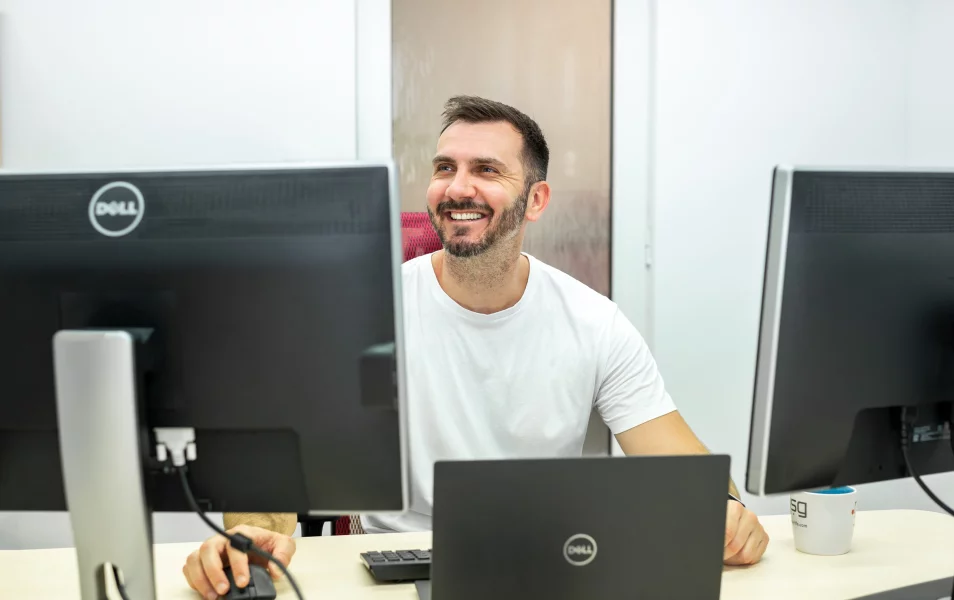 IT tester automatizovaného testovania pozerá s úsmevon nad dva monitori a latop pred ním