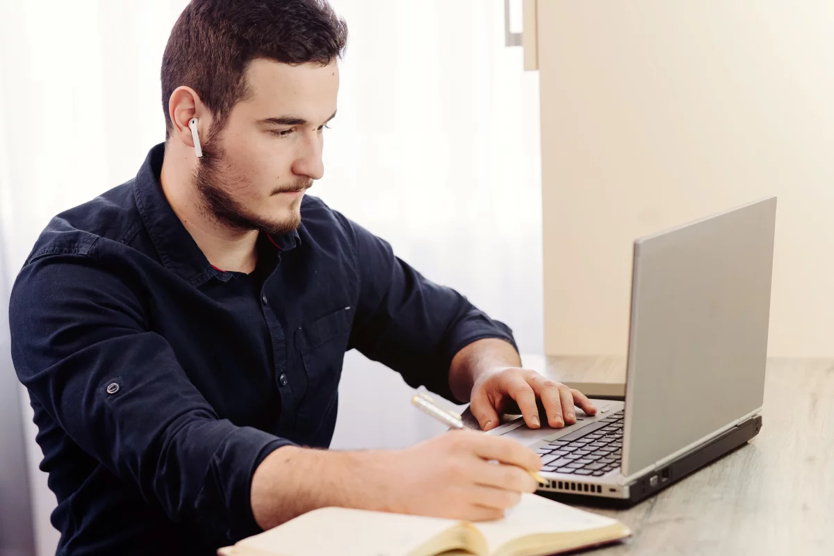 mladý muž s tmavými vlasmi v čiernej košeli pracuje na laptope a píše do poznámkového bloku